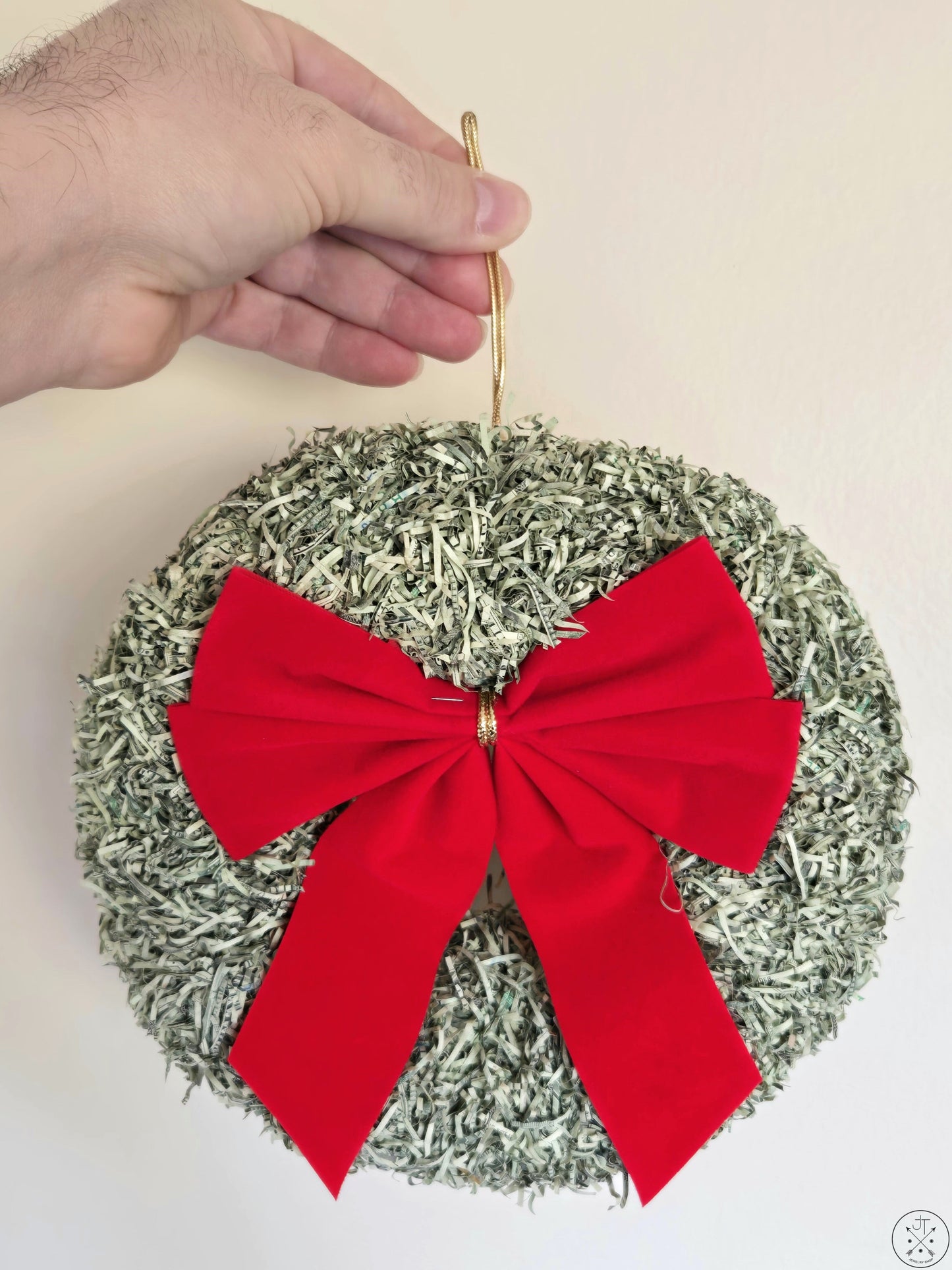 A hand holding a round Christmas wreath decoration made of shredded money with a red bow and a gold-colored ribbon.