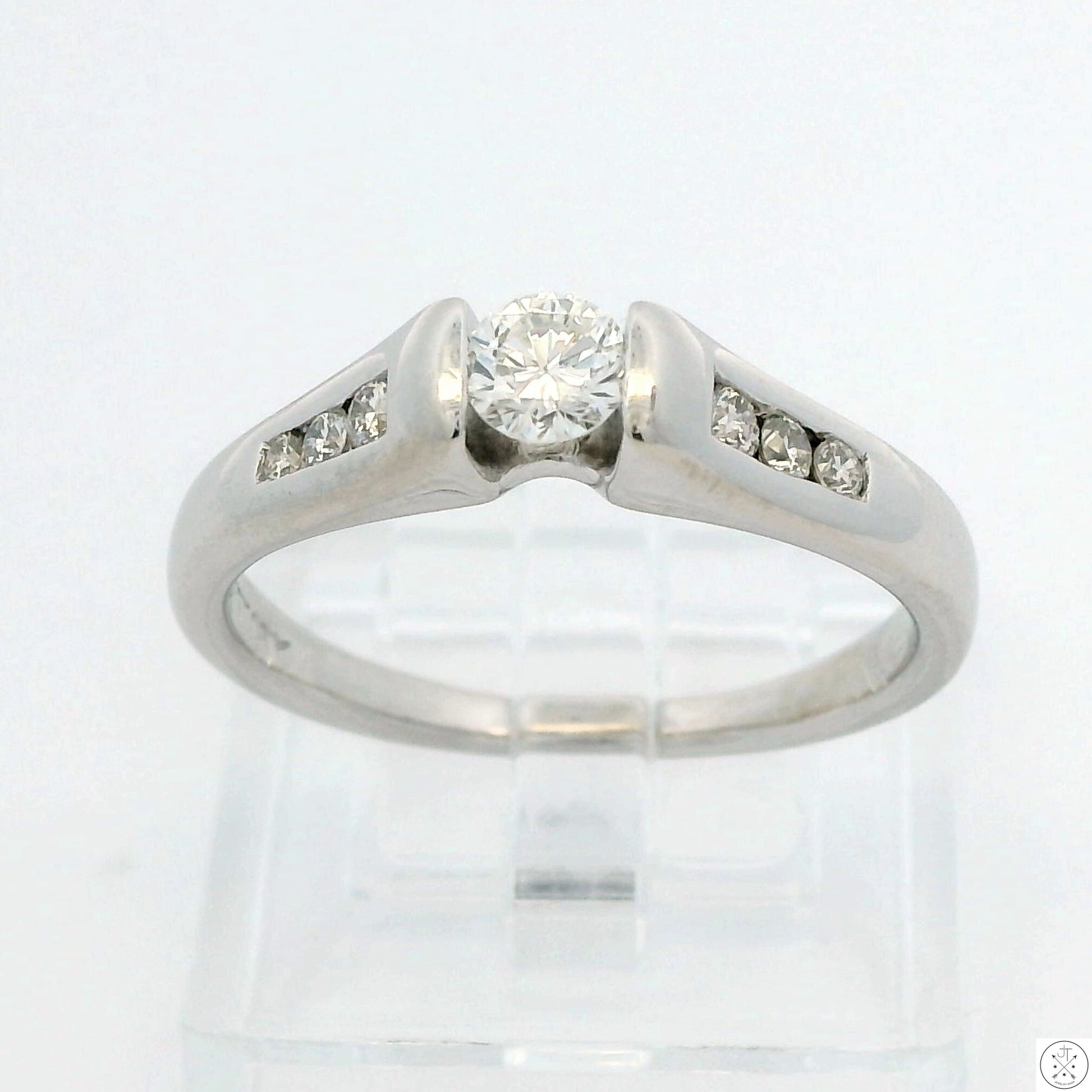 A 14k white gold engagement ring with a central diamond solitaire and accent diamonds on the band, size 7.5, displayed on a clear stand.