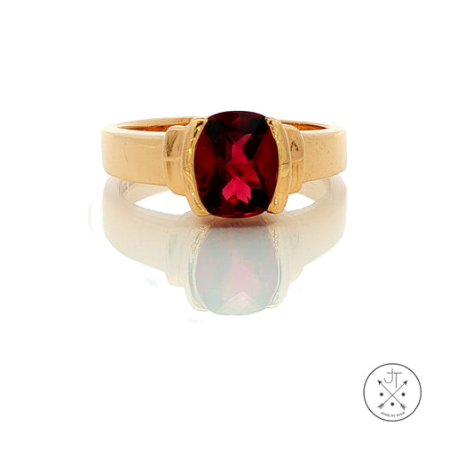 A gold ring with a large oval-cut deep red gemstone, displayed on a reflective white surface with a logo watermark in the bottom right corner.