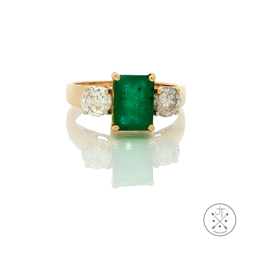 A yellow gold ring featuring a rectangular emerald-cut green gemstone in the center, flanked by two round diamonds on a white reflective background.