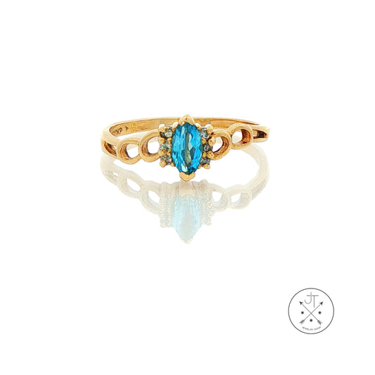 A yellow gold ring featuring a marquise-cut blue topaz center stone and small round diamonds along the shoulders, displayed on a reflective white surface.