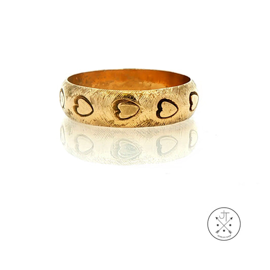 A textured gold band with engraved heart motifs, displayed on a reflective white surface with a small circular logo in the bottom right corner.