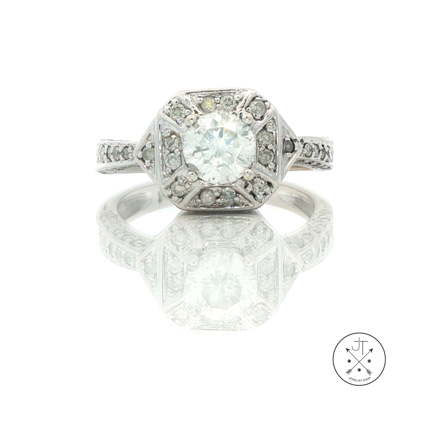 An Art Deco style diamond ring in white gold, featuring a large round center stone surrounded by smaller diamonds in a geometric setting. The ring is displayed on a reflective white surface with a logo watermark in the bottom right corner.