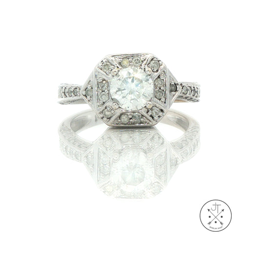 An Art Deco style diamond ring in white gold, featuring a large round center stone surrounded by smaller diamonds in a geometric setting. The ring is displayed on a reflective white surface with a logo watermark in the bottom right corner.