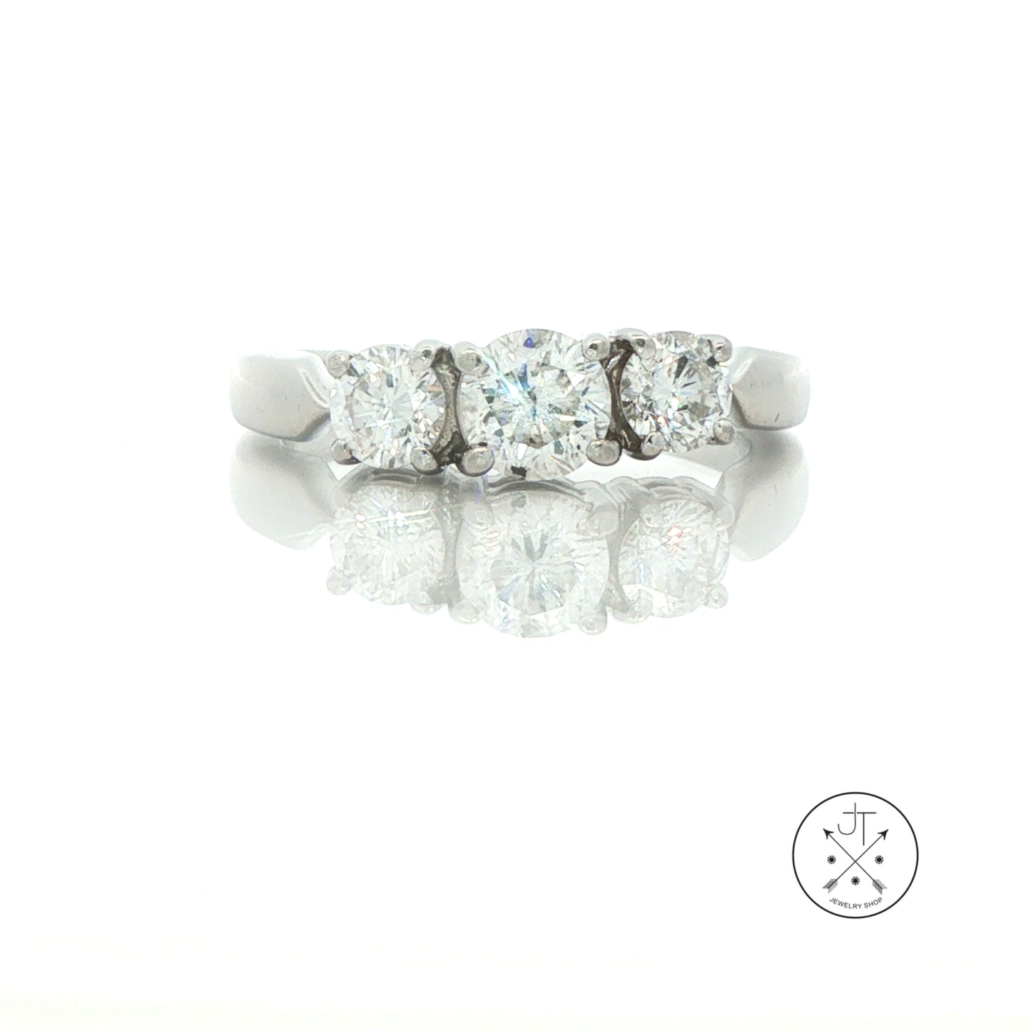 A three-stone diamond ring in a silver-colored platinum metal band, shown on a reflective white surface with a faint logo watermark in the bottom right corner.