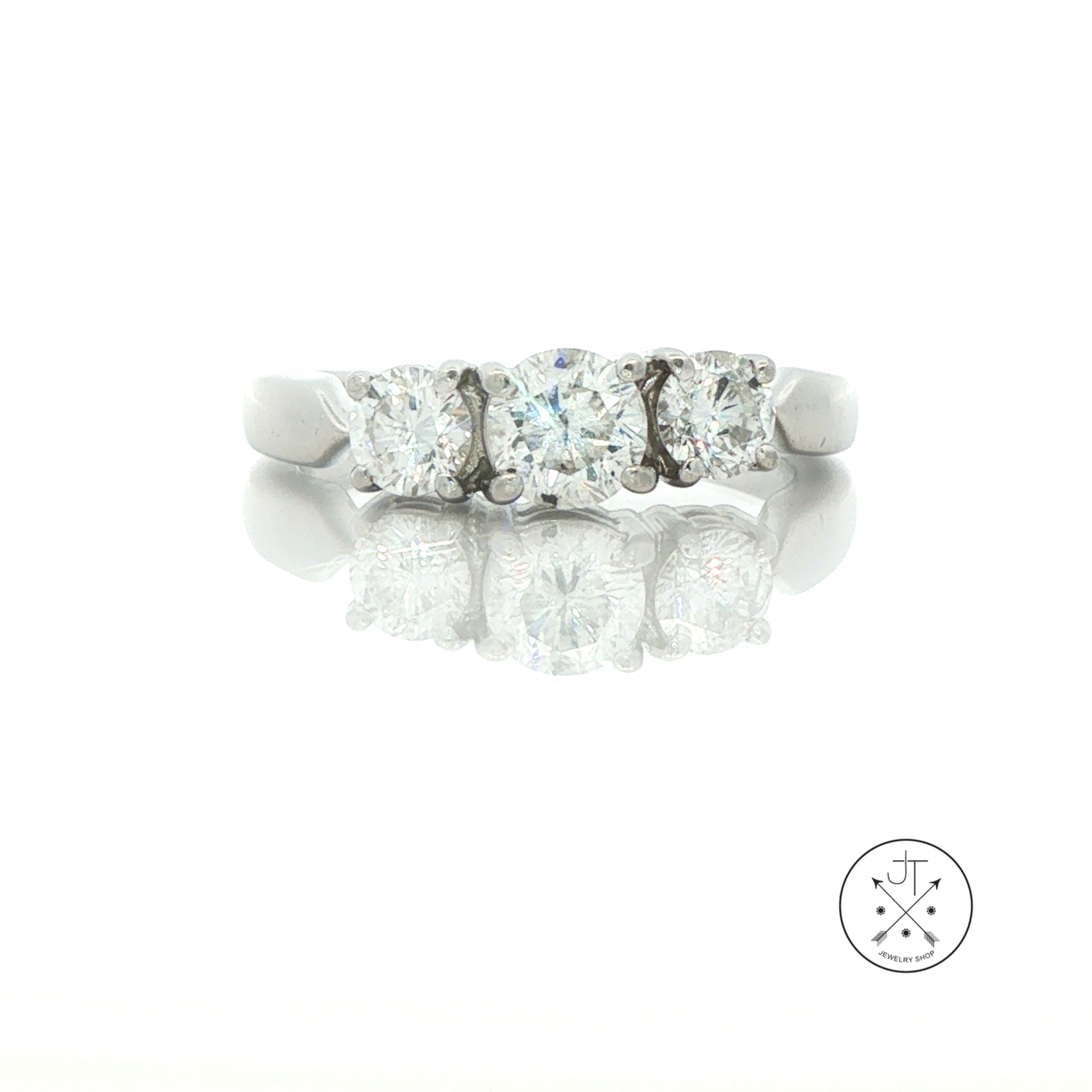 A three-stone diamond ring in a silver-colored platinum metal band, shown on a reflective white surface with a faint logo watermark in the bottom right corner.
