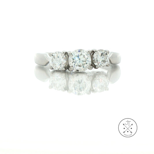 A three-stone diamond ring in a silver-colored platinum metal band, shown on a reflective white surface with a faint logo watermark in the bottom right corner.
