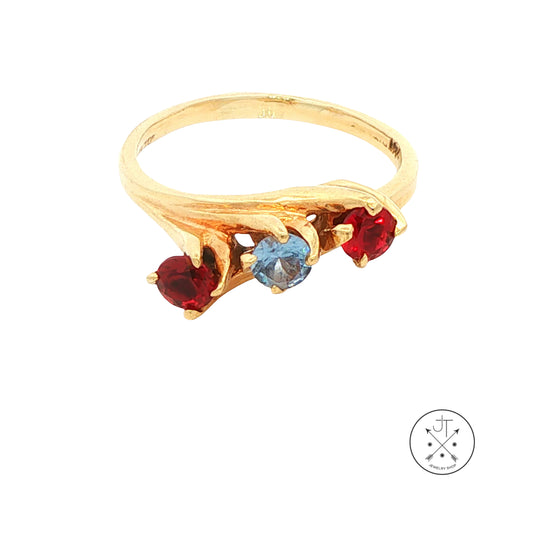 A gold ring featuring three gemstones: two round red stones and one round light blue stone, set against a plain white background with a small circular logo in the bottom right corner.