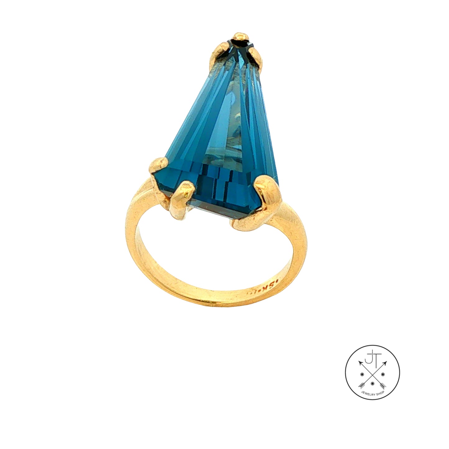 A gold ring featuring a large, faceted custom-shaped blue Topaz in a prong setting, shown against a plain white background with a small circular logo in the bottom right corner.