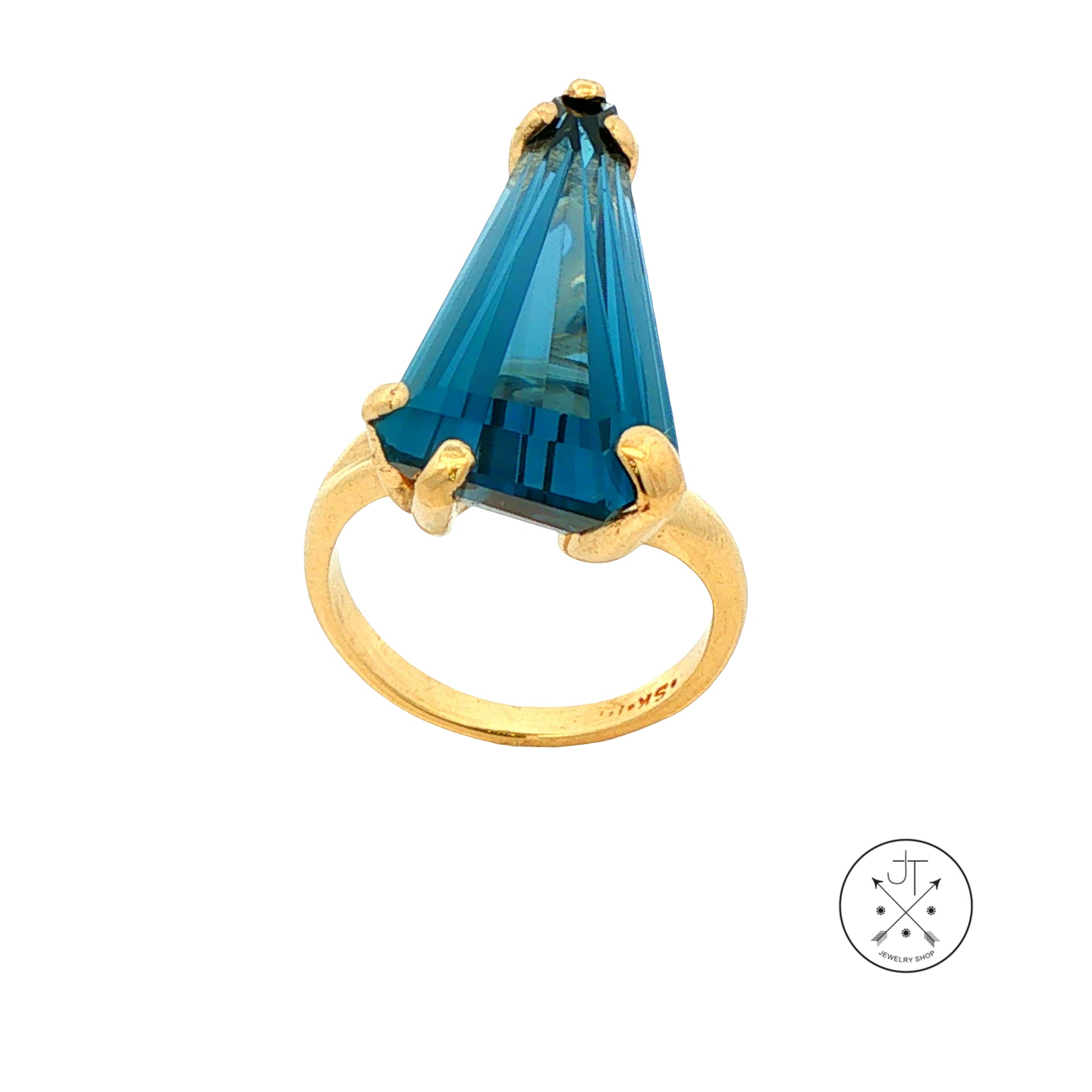 A gold ring featuring a large, faceted custom-shaped blue Topaz in a prong setting, shown against a plain white background with a small circular logo in the bottom right corner.