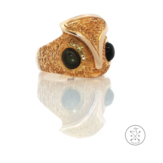 A textured gold owl ring with two large black gemstone eyes, displayed on a reflective white surface with a logo watermark in the bottom right corner.