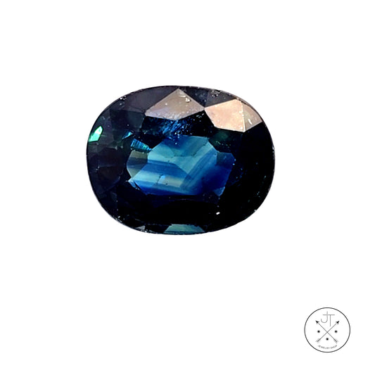 A faceted oval Sapphire gemstone with a deep blue color identity, showing iridescent blue and green reflections, isolated on a white background with a small logo in the bottom right corner.