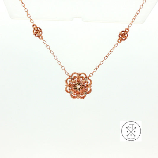 A rose gold necklace with a central floral pendant and matching decorative accents on the chain, displayed on a clean white background with a brand logo in the bottom right corner.