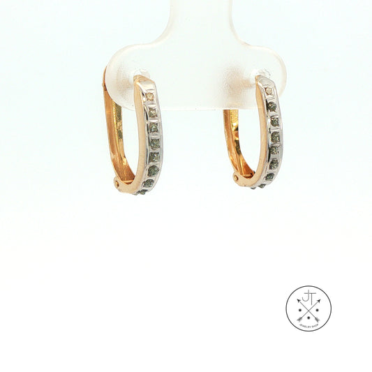 A pair of gold hoop earrings with a row of small clear stones on the front, displayed on a white stand against a white background. A circular logo is visible in the bottom right corner.